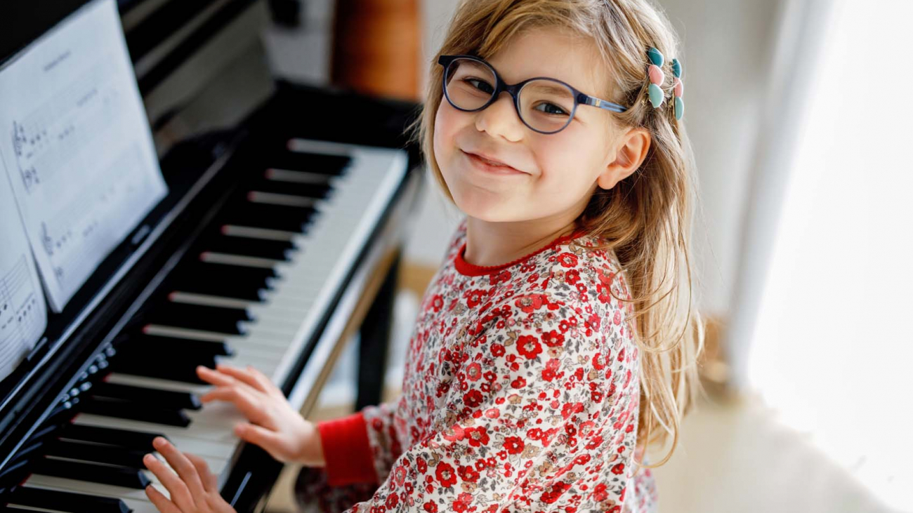 Little happy girl playing piano in living room. Cute preschool child with eye glasses having fun with learning to play music instrument