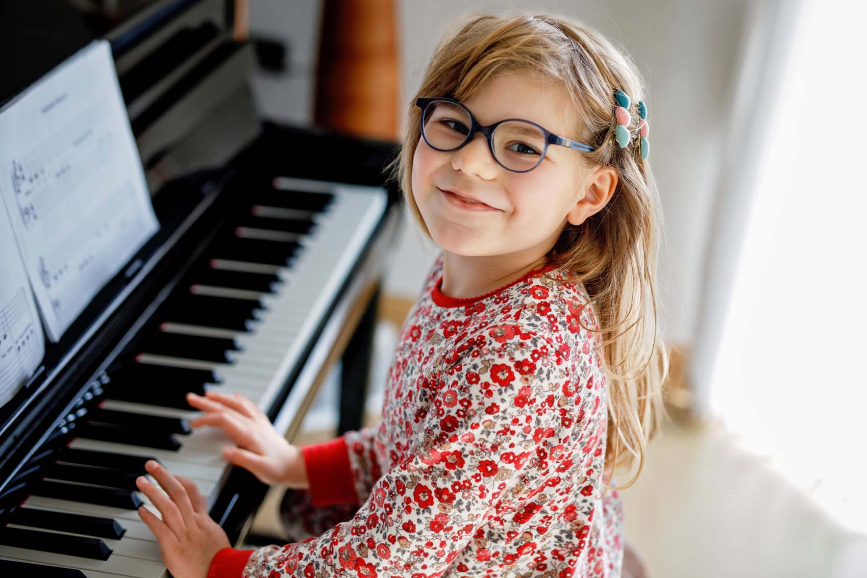 Little happy girl playing piano in living room. Cute preschool child with eye glasses having fun with learning to play music instrument
